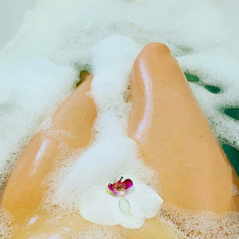 A sensual bath scene — a woman’s body submerged in soft, silky bubbles with a white orchid resting on her skin. Bath Slut’s Luxury Bubble Bath Gift Set transforms bathing into an indulgent, plant-based ritual for radiant, touchable skin.