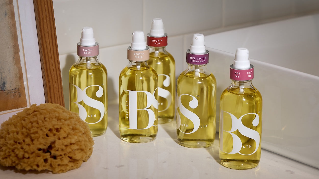 Group shot of all five Bath Slut body oils on a bathroom counter beside a sink.