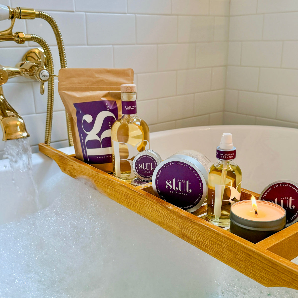 Bath Slut Ultimate Sexy Self-Care Collection in Delicious Torment, with notes of nectarine, honeysuckle and peony, on a wooden bath caddy over a bubble bath, with a lit soy candle